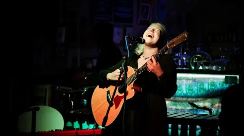 Gideon Liddiard Athena Aperta sings on stage with guitar in hand. The room is dark behind him and his face and guitar are illuminated with light. 