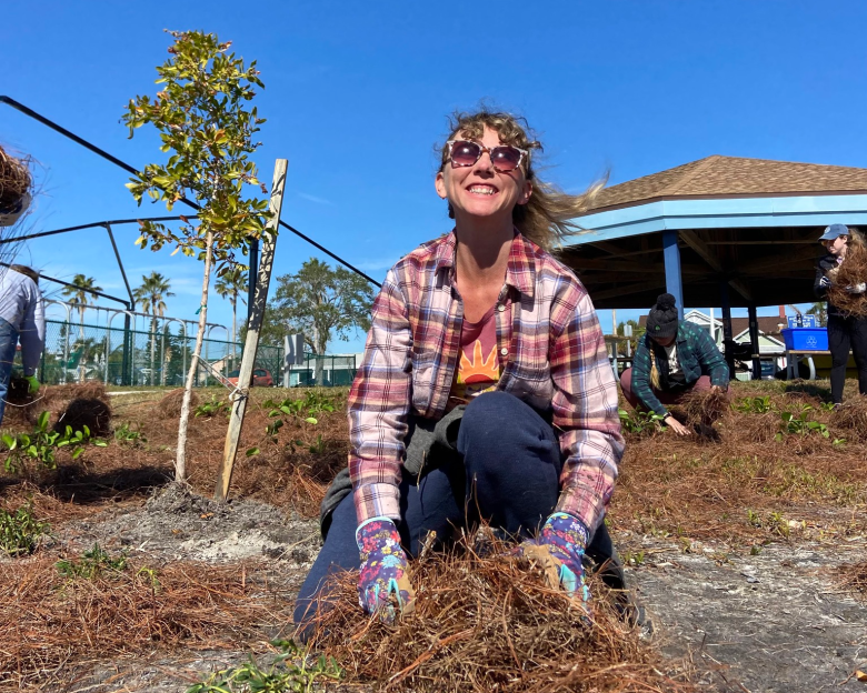 A bright outdoor daytime photo of Kira Barrera volunteering at a community tree planting event with the Florida Suncoast Sierra Club. In the center foreground, they squat in a large pile of brown pine straw mulch, looking up and smiling warmly. They wear checkered flannel shirts in red, white, and navy blue over dark tops, dark pants, sunglasses, and work gloves with colorful patterns. In the background on the left, a newly planted slender sapling stands supported on stakes under a clear, bright blue sky. On the right you can see an outdoor park pavilion with a blue-trimmed roof, and in the background you can see other people doing volunteer work.