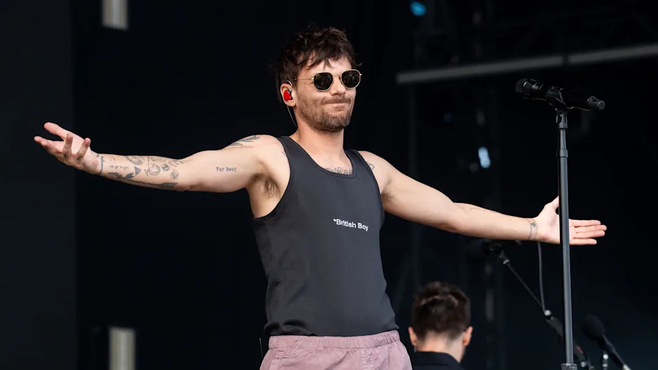 Louis Tomlinson performs on stage wearing a black T-shirt "british boy" It's written there. He also wears sunglasses and pale pink pants. 