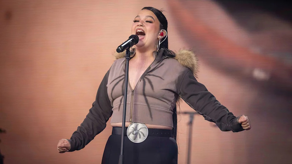 Laura Young is singing into a microphone on stage. She wears a brown zip-up jacket with a fur collar hood and black pants. 
