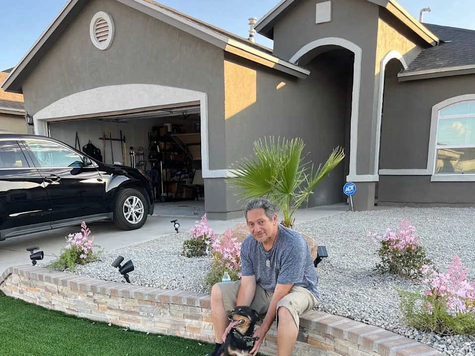 A man is sitting in front of his house with a small dog in his arms.
