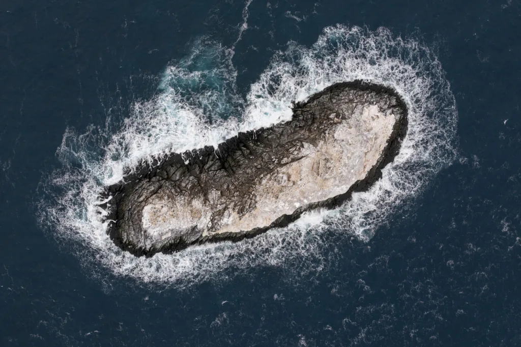 Aerial view of a small rocky island surrounded by waves crashing in a deep blue sea.
