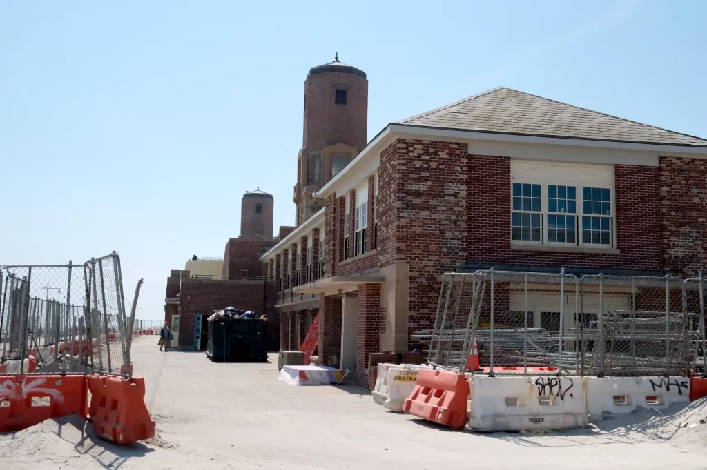 Construction is underway on the historic Jacob Riis Bathhouse. It features a brick building and two towers, with a fence and building materials in the foreground.